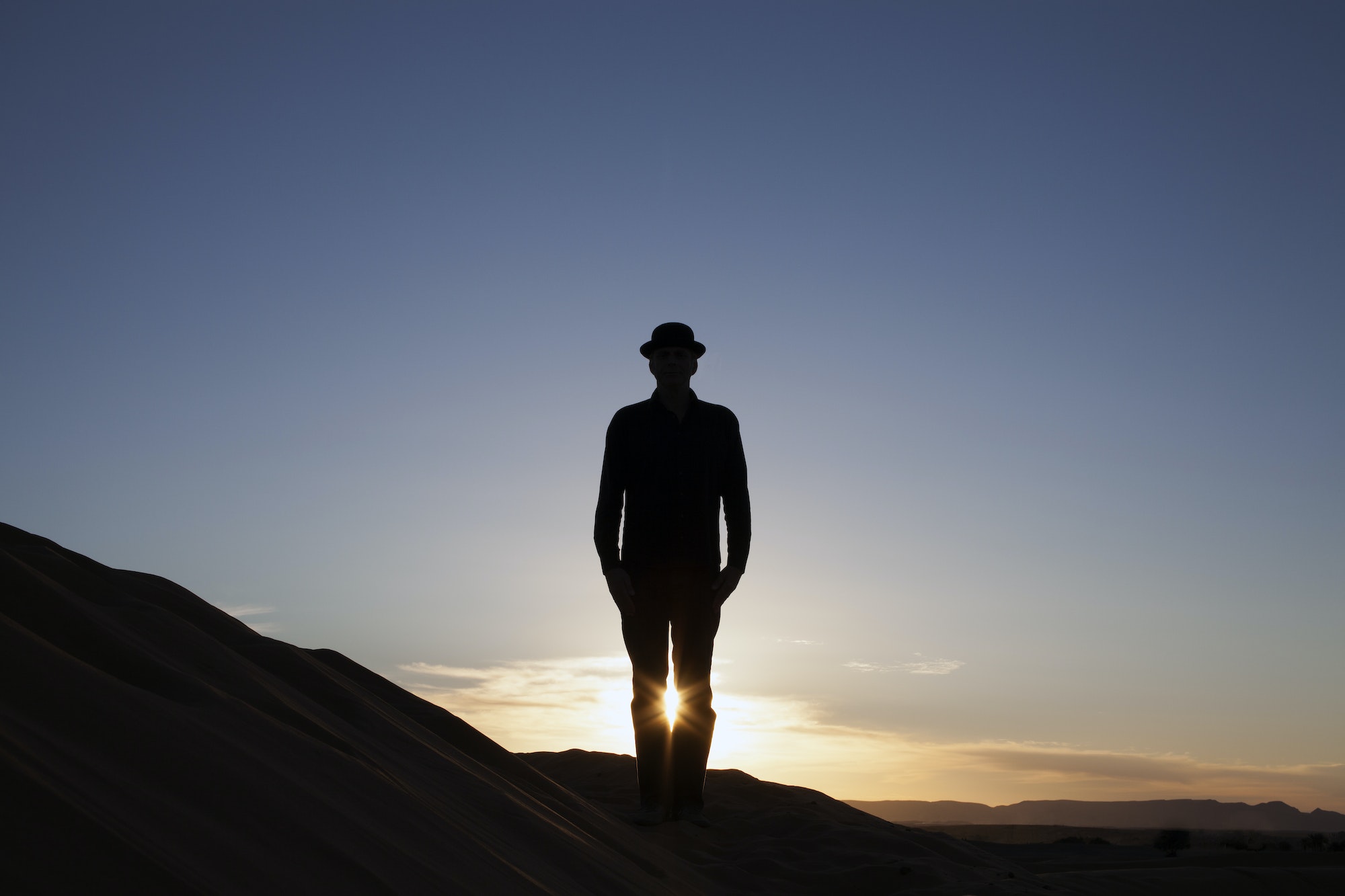 Morocco, Merzouga, Erg Chebbi, silhouette of man wearing a bowler hat standing on desert dune at sun, 9 days to Desert from casablanca