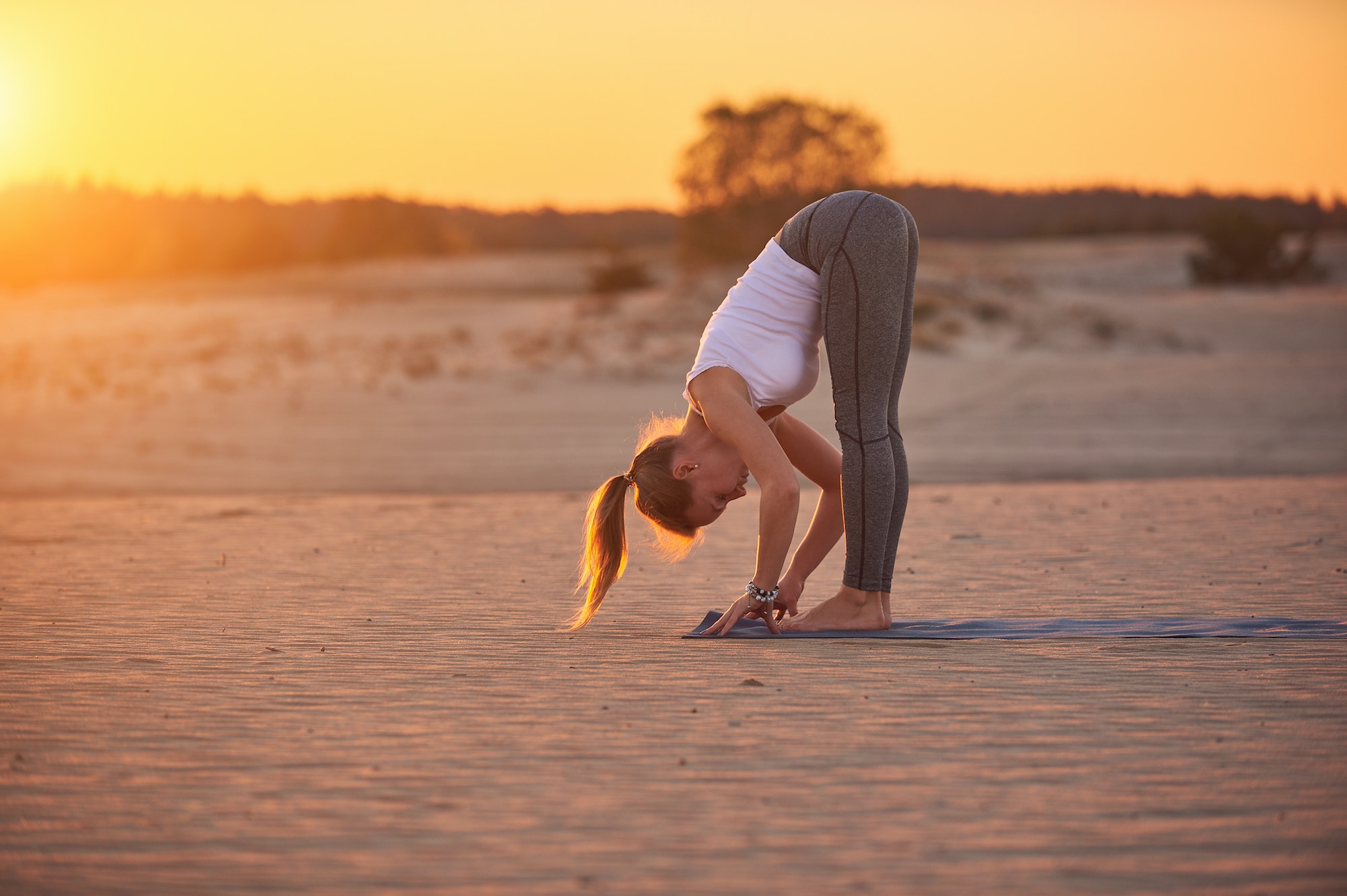 Young woman is doing yoga asana Uttanasana - Standing Forward Fold in the desert at sunset
