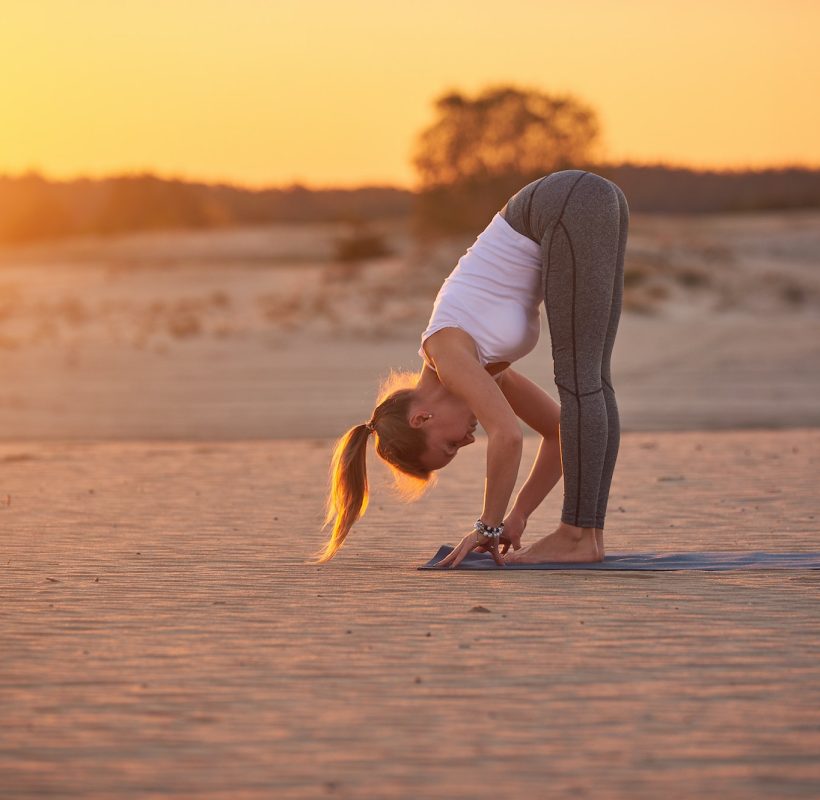 Young woman is doing yoga asana Uttanasana - Standing Forward Fold in the desert at sunset